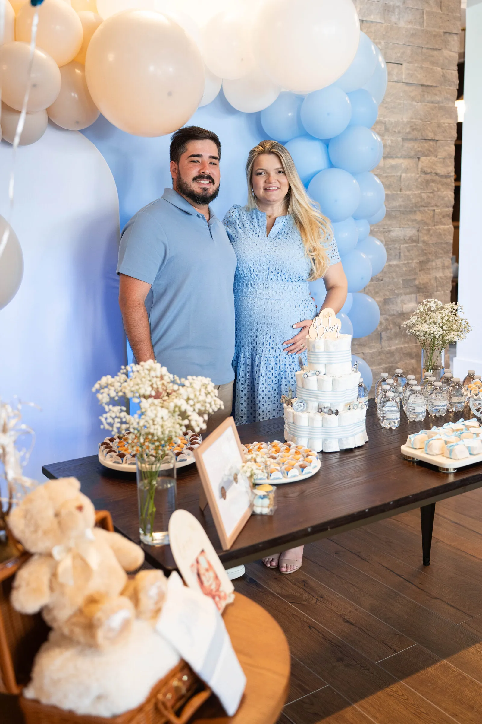 Expecting parents standing arm in arm in front of a balloon arch and dessert table