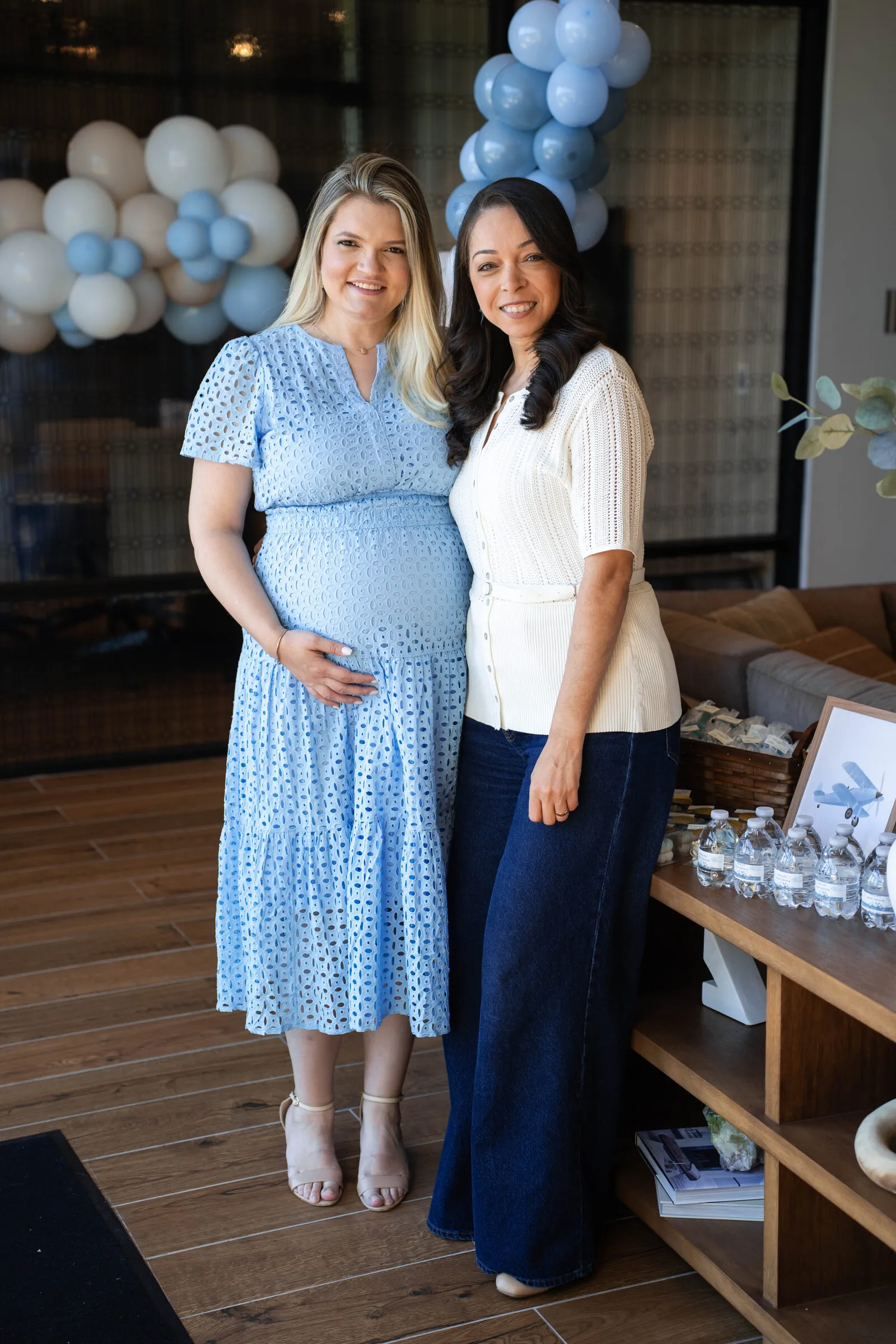 Two guests standing arm in arm in front of a blue and cream balloon garland at the baby shower