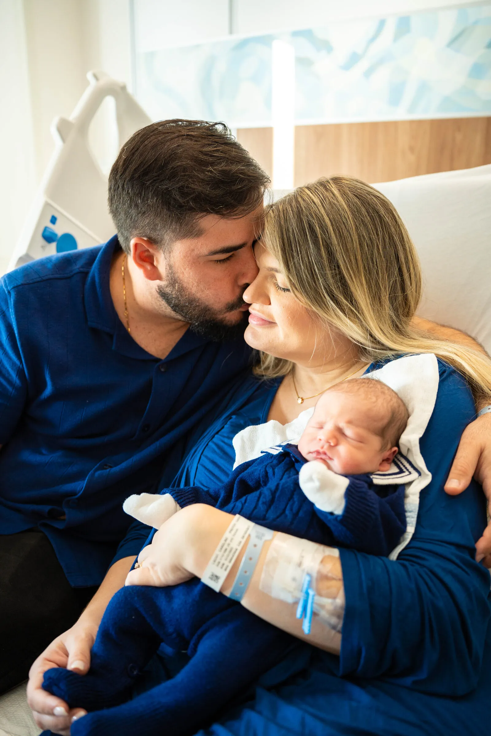 A father kisses the mother's head while she holds their newborn in the maternity room