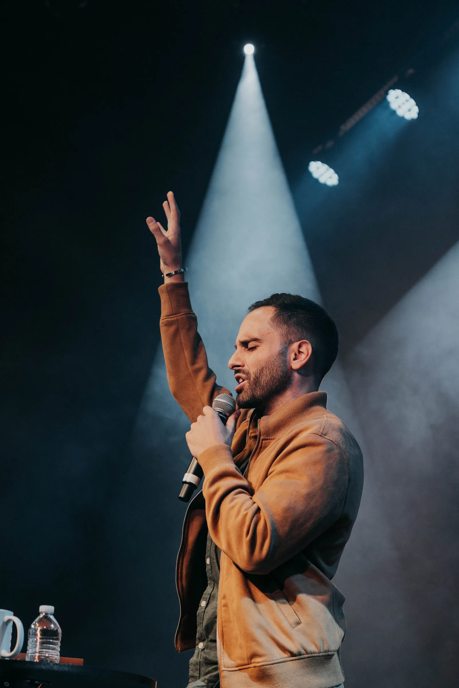 Singer with arms raised in a hard spotlight beam during a Poiema worship night